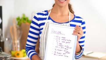 Woman in the kitchen at home, standing near stove while holding a recipe.