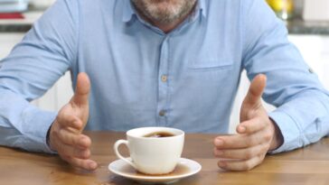 Unidentifiable man brewing coffee at home spills his coffee, looking at camera with sad and confused expression.