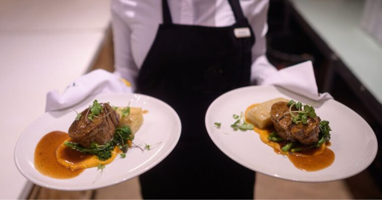 Waiter holding hot plated grilled steak and vegetables potatoes asparagus butter nut garnish sauce.