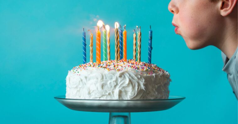Boy blowing out candles on a birthday cake against blue background.