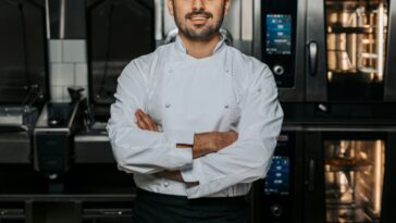 Portrait of confident, young male chef wearing a white uniform and standing with his arms crossed in the kitchen of a restaurant.