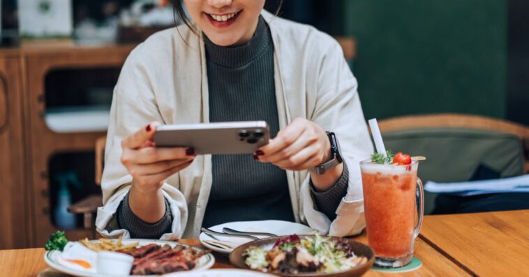 Smiling young woman taking photos of her scrumptious and beautifully plated dishes on the table with her smartphone in outdoor cafe. Enjoying her meal and sharing foodie photos on social media. Digital dining. People, food, lifestyle and technology.