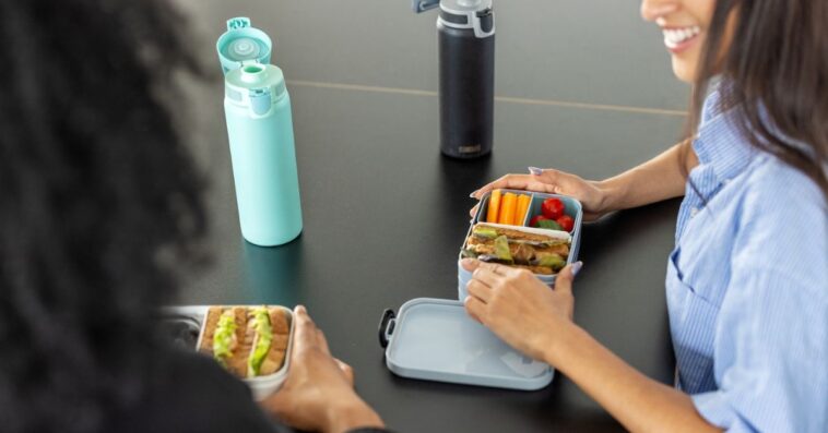 Close-up of two woman coworkers sitting at desk with lunch boxes in office. Healthy food in lunch boxes for female colleagues at work desk.