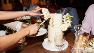 Cropped hands of newlywed couple cutting wedding cake near guests sitting at illuminated dining table during reception dinner party.