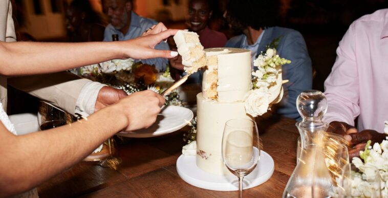 Cropped hands of newlywed couple cutting wedding cake near guests sitting at illuminated dining table during reception dinner party.