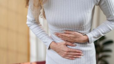 Close-up of a woman's midsection. She is wearing a white dress and is holding her stomach in pain.