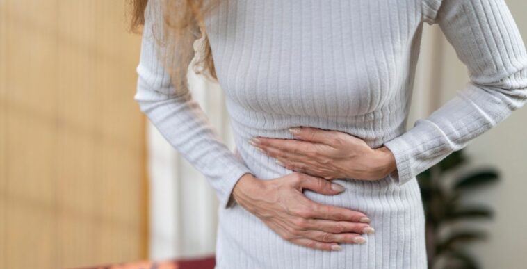 Close-up of a woman's midsection. She is wearing a white dress and is holding her stomach in pain.