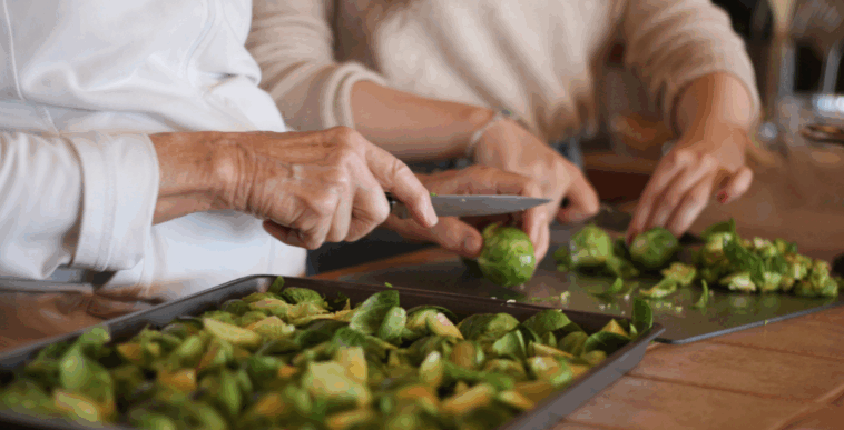 Two women cutting up brussell sprouts at a kitchen counter.