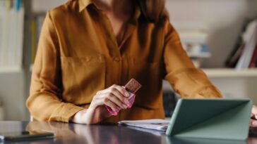 An anonymous businesswoman sitting at the office, eating a protein bar while video calling with her colleagues.