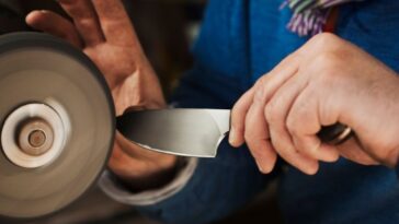 Close up of a craftsmans hands, holding a strong steel knife blade against a surface grinder, hand finishing a kitchen knife.