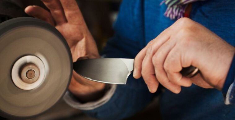 Close up of a craftsmans hands, holding a strong steel knife blade against a surface grinder, hand finishing a kitchen knife.
