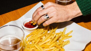 A woman's hand with rings, dips fries in ketchup and their is a beer on the table.