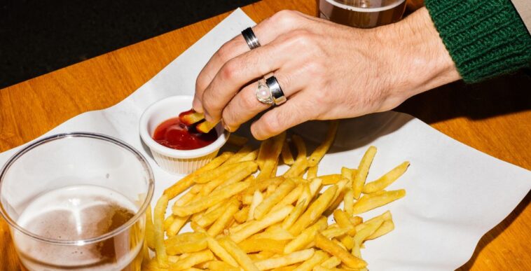 A woman's hand with rings, dips fries in ketchup and their is a beer on the table.