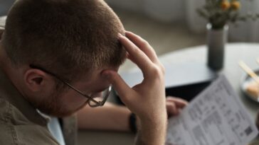 A young adult man sits at a table holding overdue bills, looking stressed and frustrated, hand on forehead, financial documents and chopsticks visible on surface.