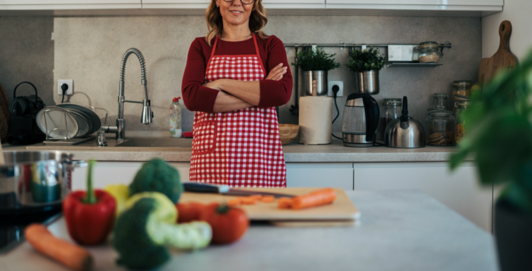 A woman wearing a red checkered apron with her arms crossed in a kitchen