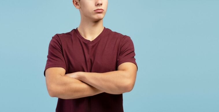 Serious, sad teen boy with crossed arms looking away on copy space, standing on blue background.