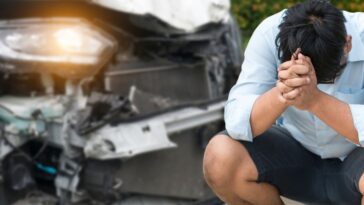 A distressed man sits on the curb in front of a car accident, he has his head in hands.