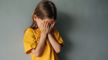 A sad, young girl cries into her hands, she stands against a gray backdrop.