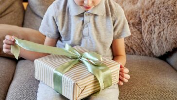 Curious young boy on a comfy sofa carefully unties a light green ribbon on a striped gift box, eagerly anticipating the surprise inside during a cozy home moment.