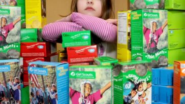 A young girl surrounded by a ton of girl scout cookie boxes looks out longingly.