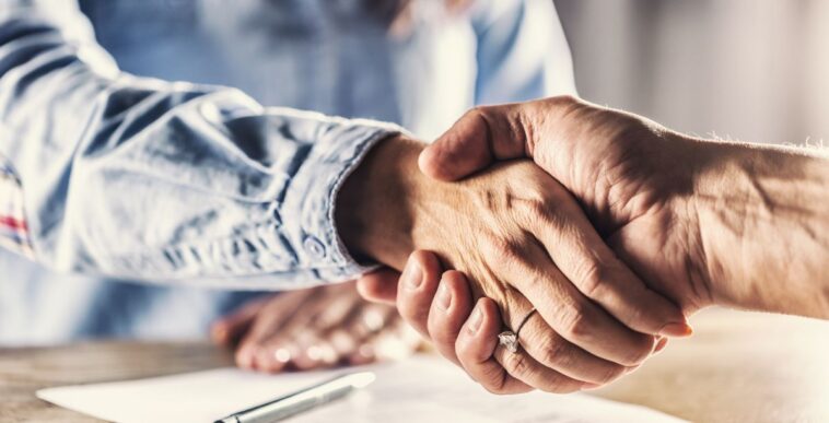 Female and male shake hands over a closed deal signed by a pen on a paper below.