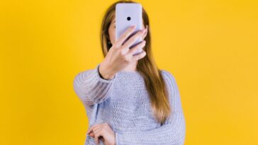Young woman holding smartphone in front of her face against a yellow backdrop.
