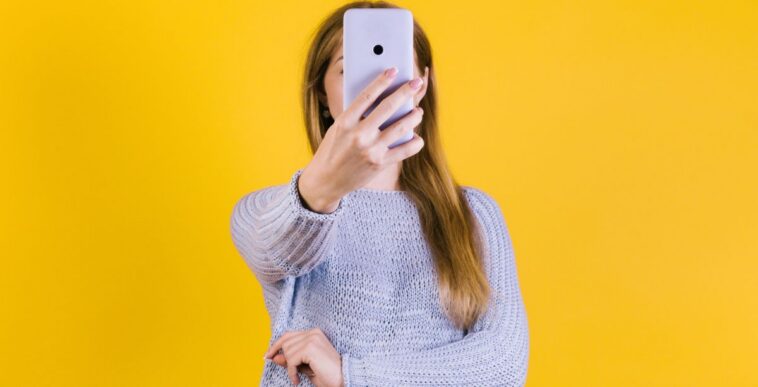 Young woman holding smartphone in front of her face against a yellow backdrop.