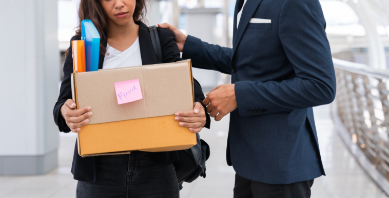 A woman holding a cardboard box full of items, with a man standing next to her resting his arm on her shoulder.