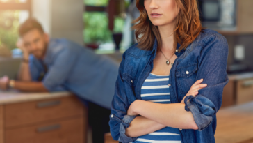 A woman with her arms folded and a man leaning against a counter in the background.
