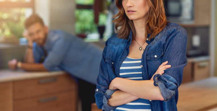 A woman with her arms folded and a man leaning against a counter in the background.