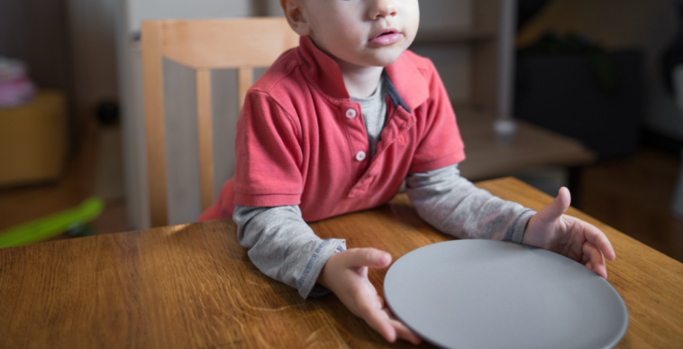 A little boy sitting at a table, holding an empty plate.