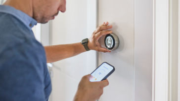 A man adjusting a thermostat on the wall and with his phone.