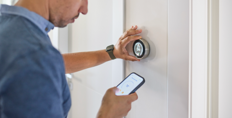 A man adjusting a thermostat on the wall and with his phone.