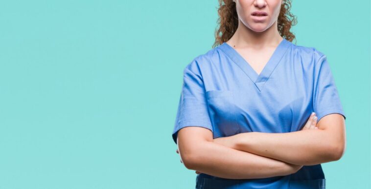 Young, brunette female doctor wearing scrubs, over isolated background skeptic and nervous, disapproving expression on face with crossed arms.