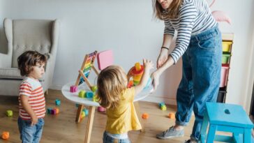 Positive young female babysitter in casual clothes playing with infant children in cozy playroom with colorful educational toys.