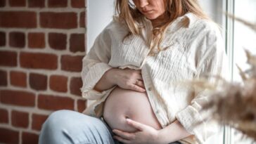 Troubled expectant mother sits on the windowsill, dressed in a muslin shirt and denim jeans.