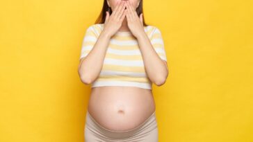 Shocked, astonished pregnant woman with bare belly wearing casual top isolated over yellow background screaming with amazement covering mouth with hands.