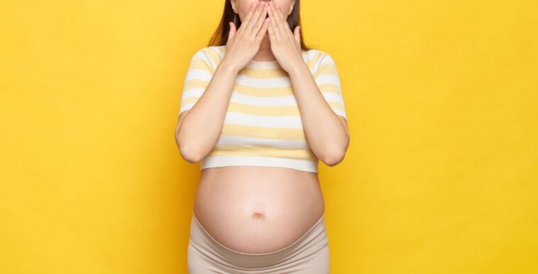 Shocked, astonished pregnant woman with bare belly wearing casual top isolated over yellow background screaming with amazement covering mouth with hands.