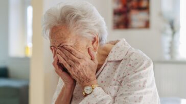 Senior woman crying. Elderly lady has headache, feeling dizzy after waking up. Elderly woman in pyjamas sitting on bed, holding her head.