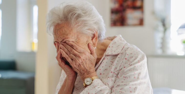 Senior woman crying. Elderly lady has headache, feeling dizzy after waking up. Elderly woman in pyjamas sitting on bed, holding her head.