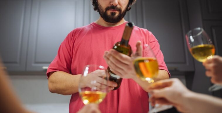 A man with a beard in a pink shirt is holding a bottle of wine while hands with wine glasses are extended to him. The background shows a kitchen scene. Suitable for gatherings, parties, and social events.