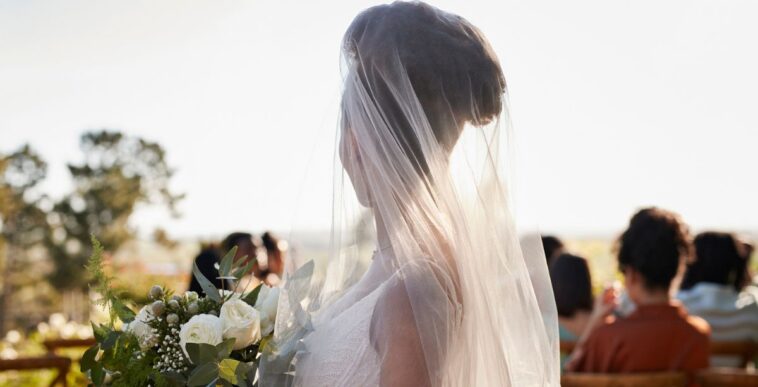 Side view of a young bride wearing a veil, standing with a flower bouquet at a wedding ceremony.